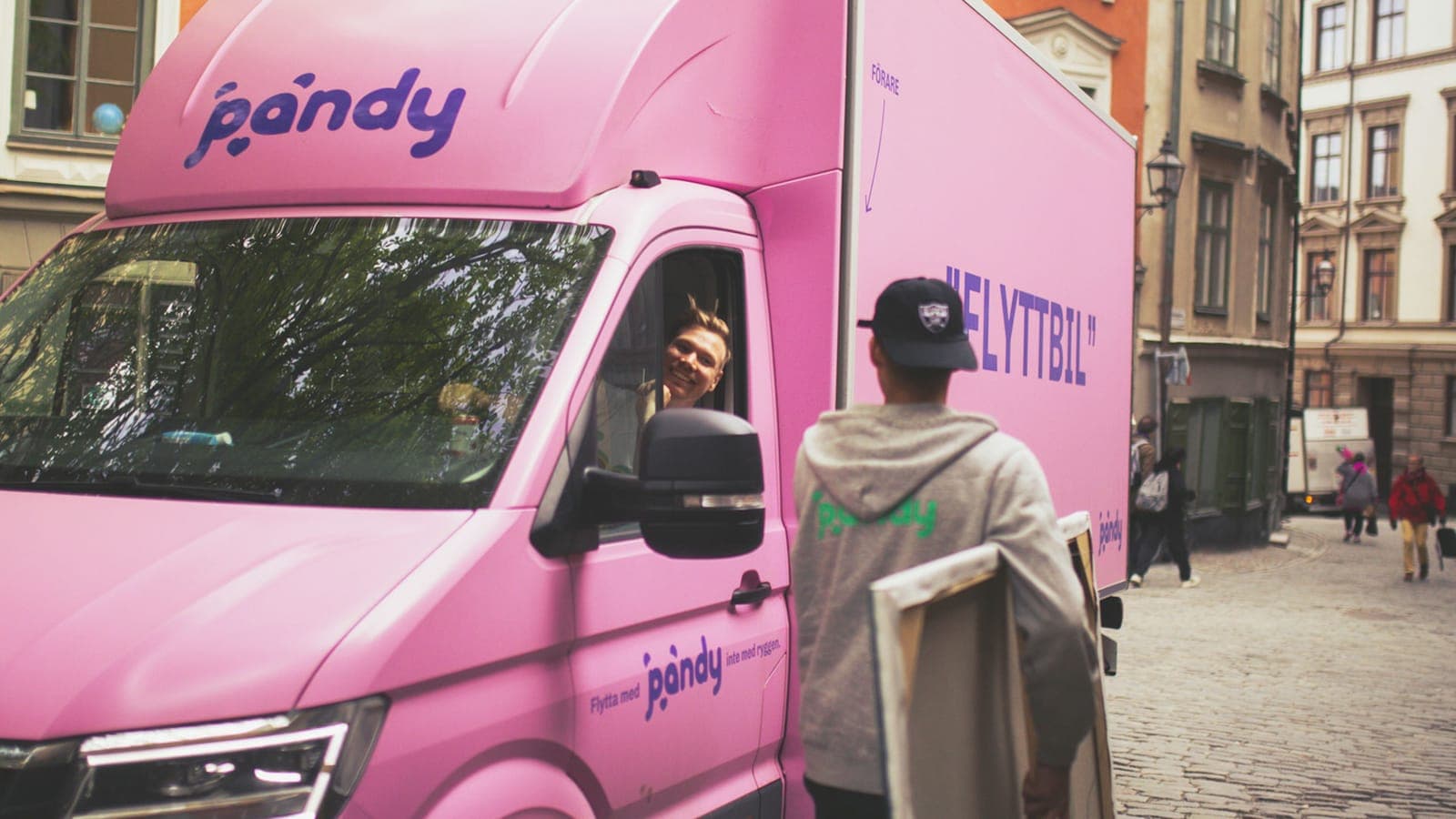 Pink moving van on a street on a sunny day, with a person in front of it carrying a picture